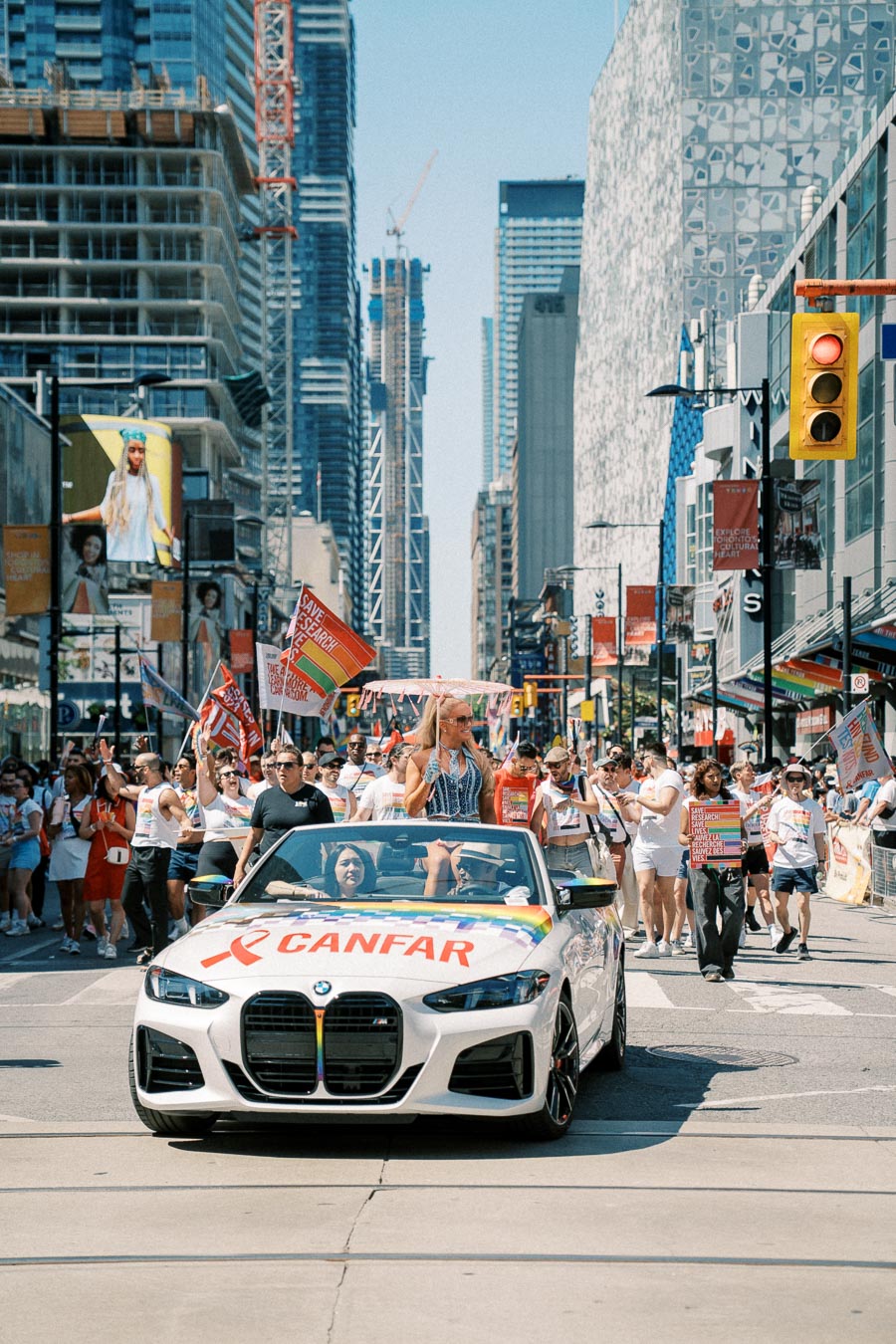 A vibrant parade scene featuring a white BMW convertible with a CANFAR banner, surrounded by participants holding flags and signs, set against a backdrop of urban skyscrapers.