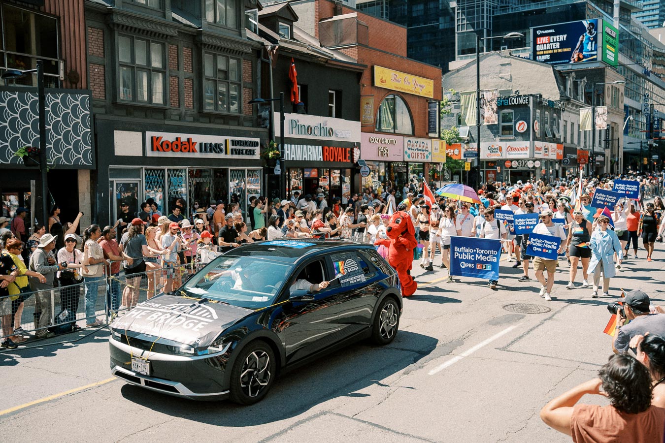 A vibrant parade takes place on a city street, featuring a black car and participants holding signs for Buffalo-Toronto Public Media. Spectators line the sidewalks, enjoying the festive atmosphere.