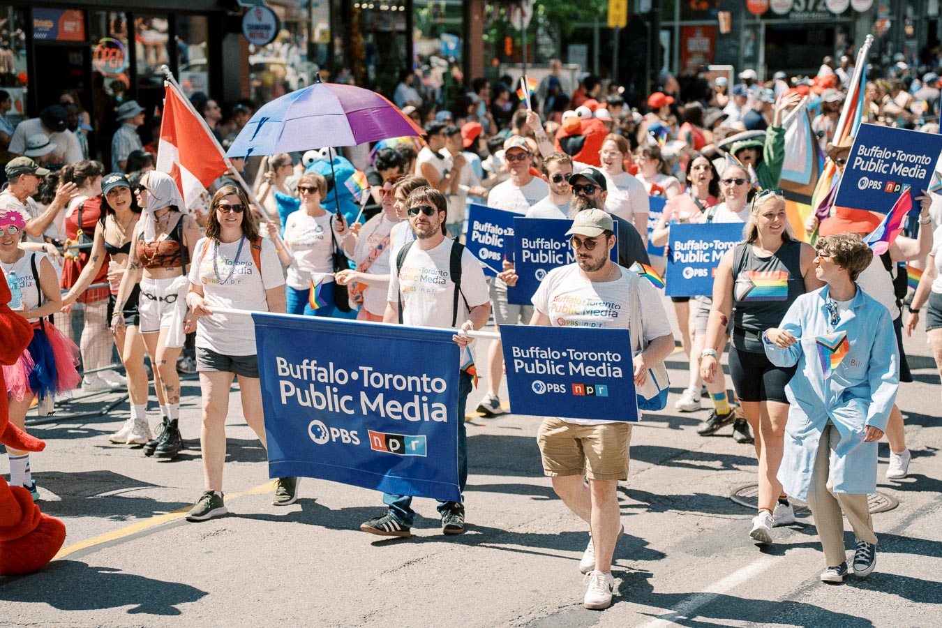 Participants in a parade hold Buffalo-Toronto Public Media signs, celebrating with rainbow flags and vibrant attire.