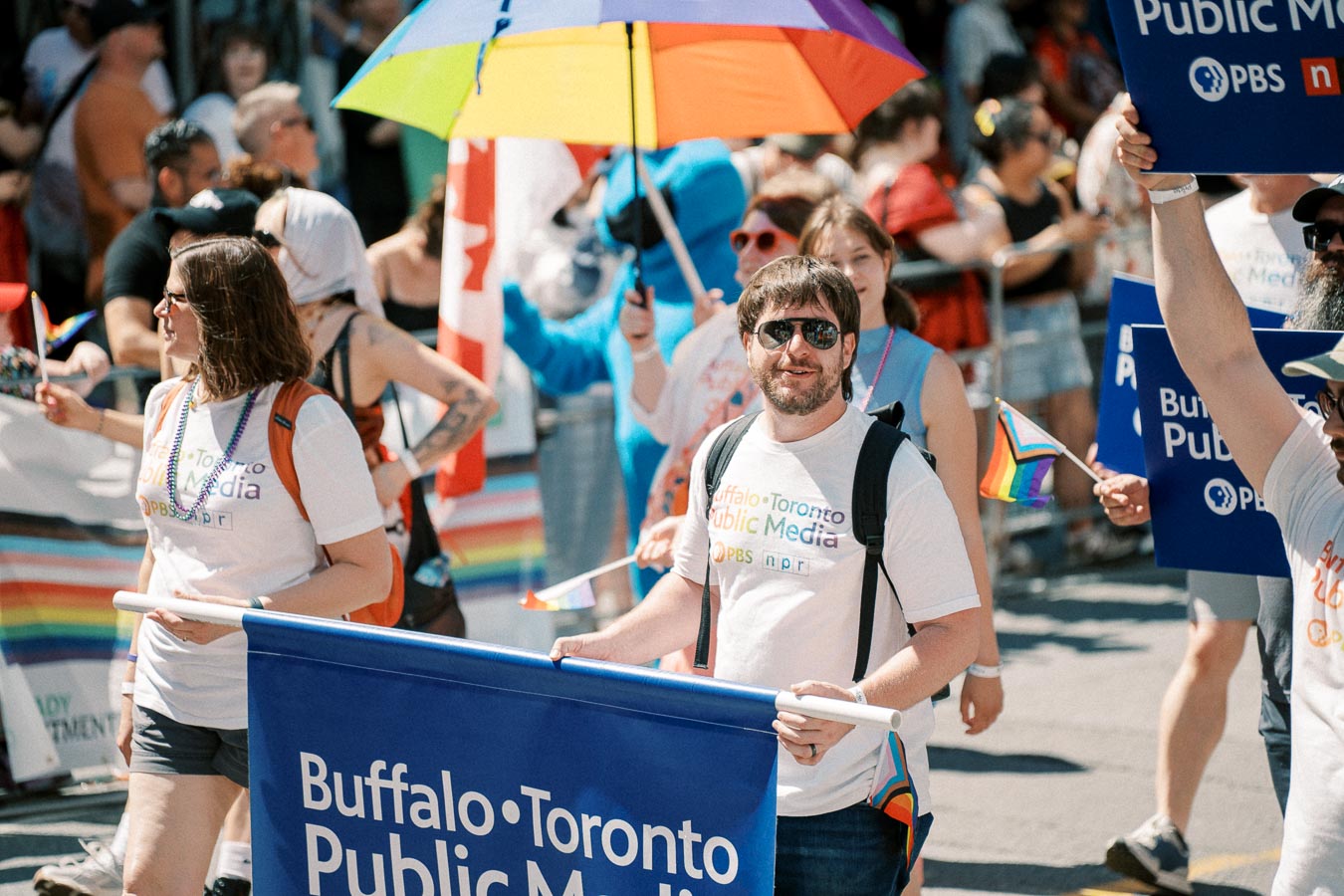Participants in a parade hold a Buffalo Toronto Public Media banner, celebrating with rainbow flags and a colorful umbrella.