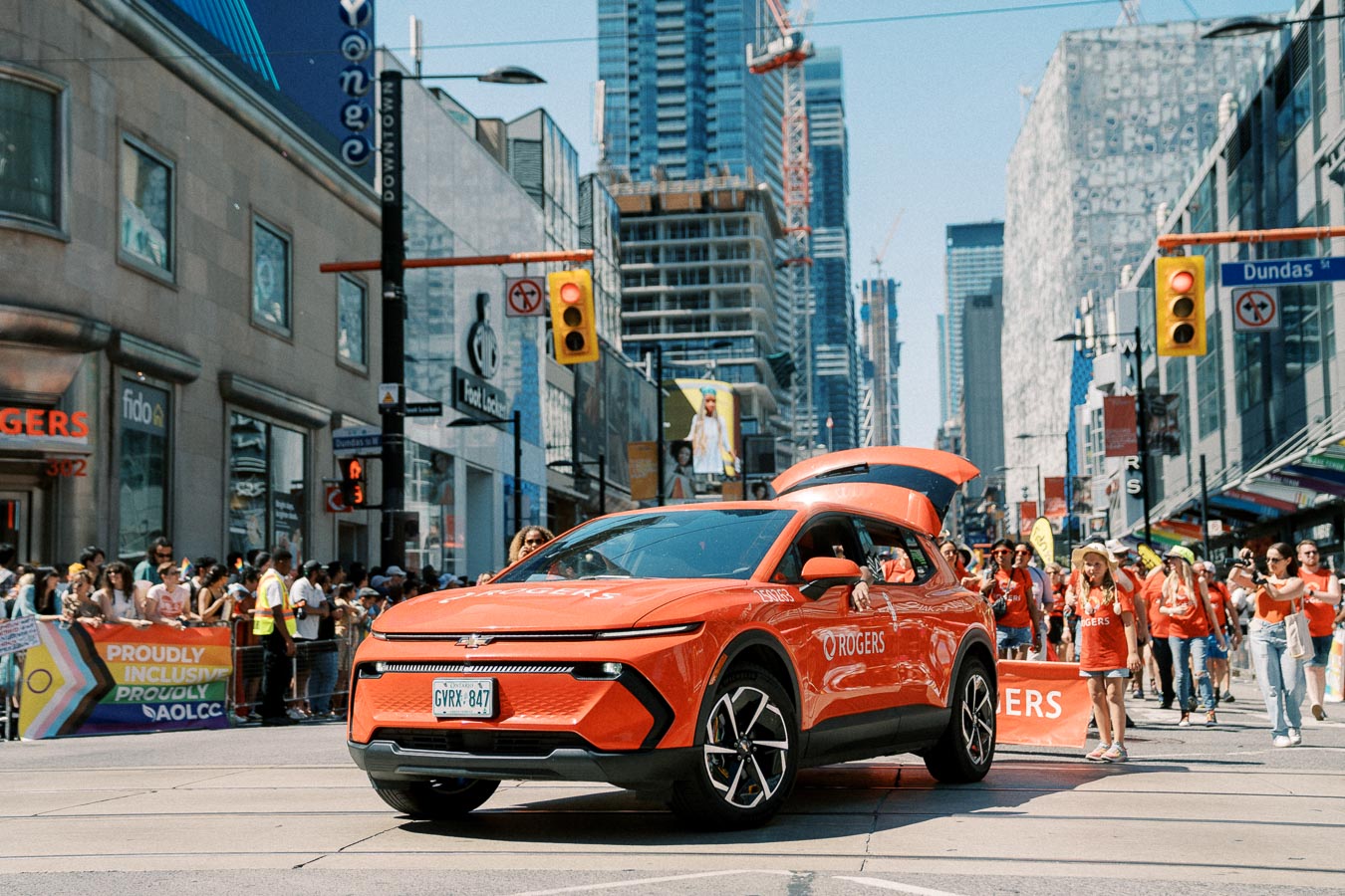 A red electric vehicle branded with Rogers is seen participating in a parade on a bustling city street, with crowds of people and tall buildings in the background.