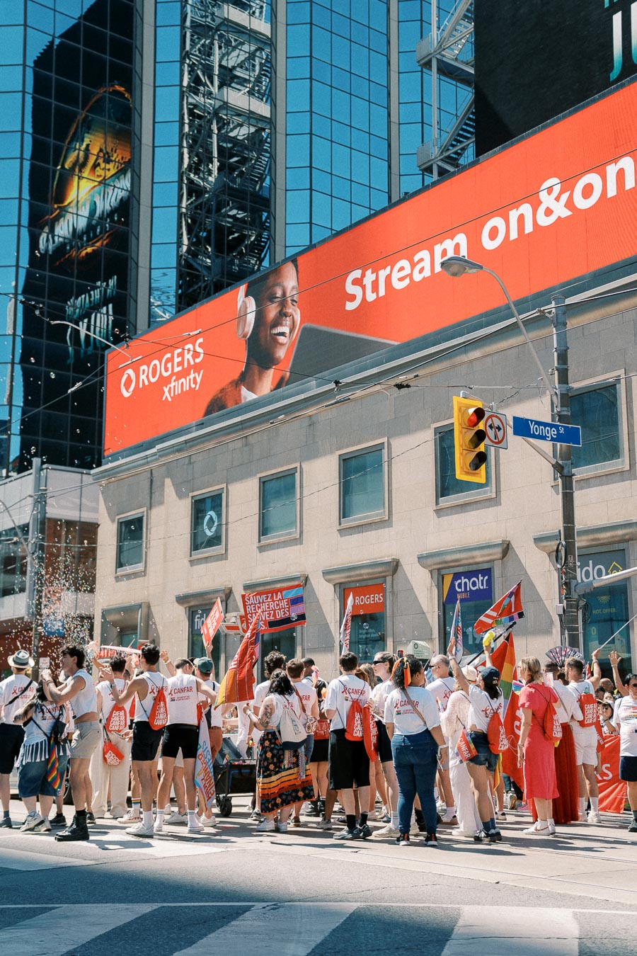 A lively gathering on Yonge Street in Toronto, with people holding flags and wearing matching shirts under a prominent Rogers Xfinity billboard.