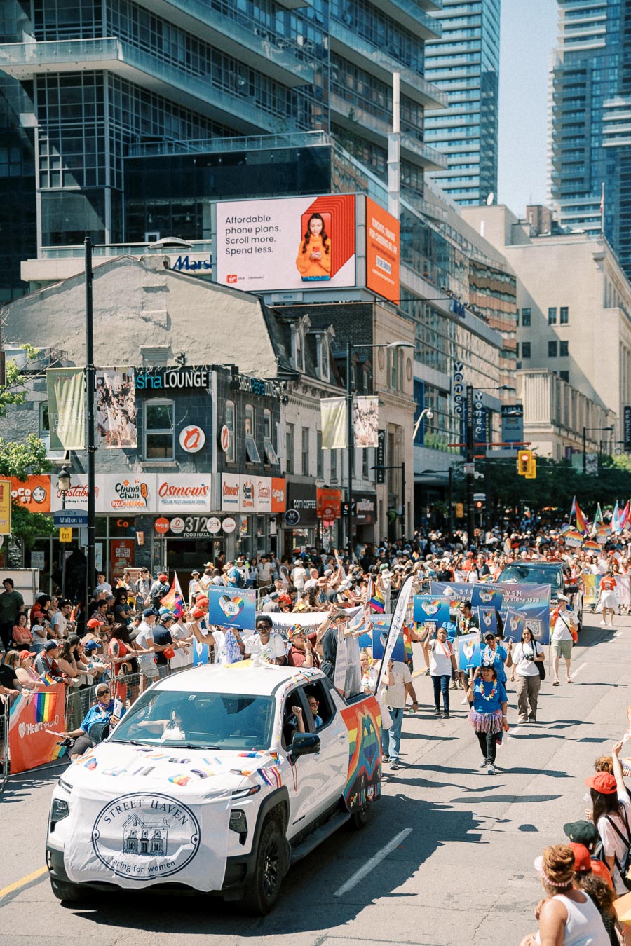 A lively parade featuring a diverse group of participants marching down a city street, with banners and flags. A decorated white truck leads the procession, set against a backdrop of skyscrapers and a digital billboard.