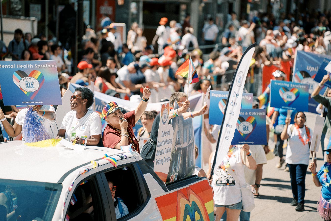 Participants in a parade celebrate diversity and mental health services, holding signs and rainbow flags.