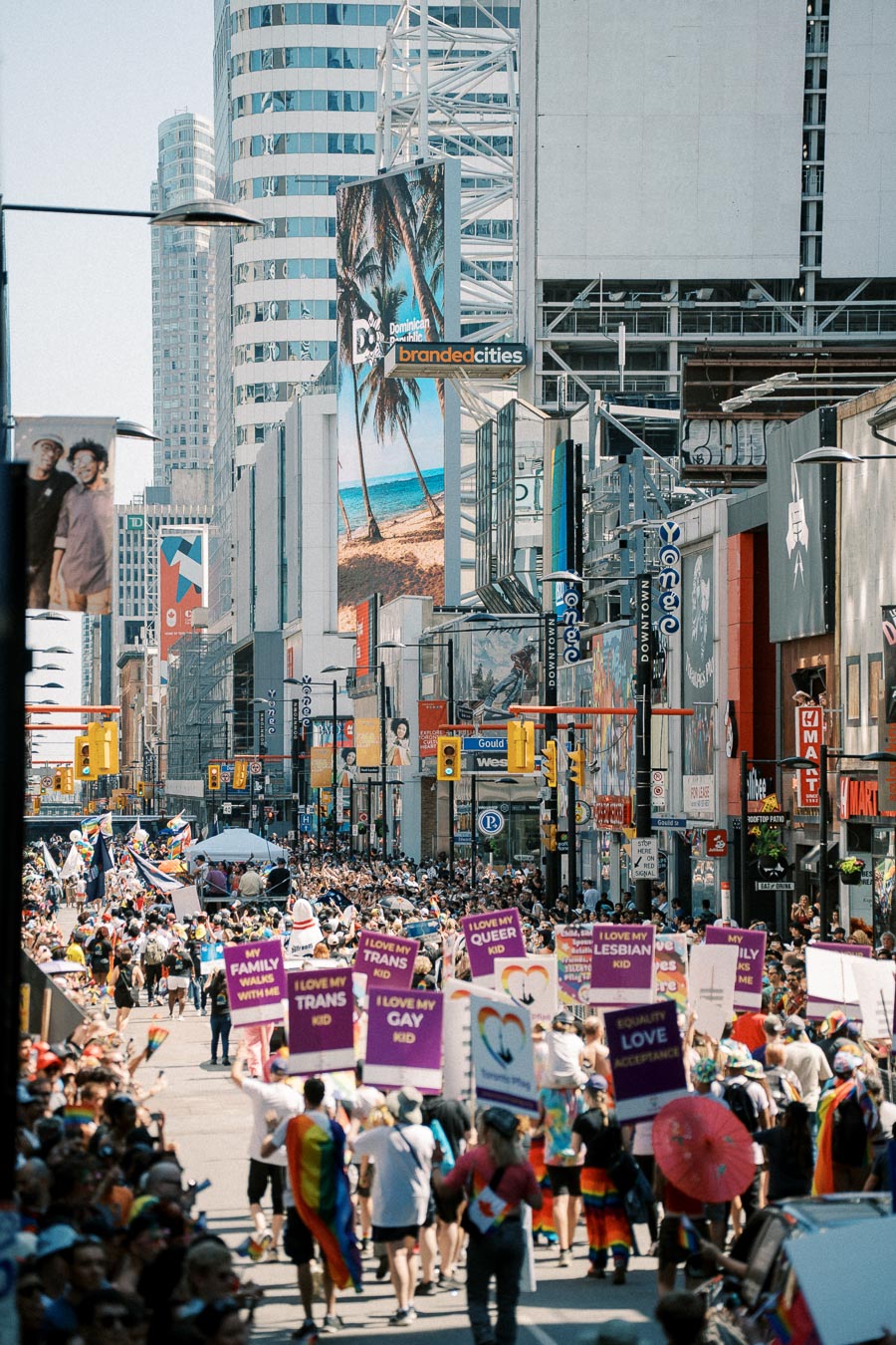 A lively pride parade takes place on a bustling city street, surrounded by skyscrapers and colorful billboards. Participants carry signs supporting LGBTQ+ rights.