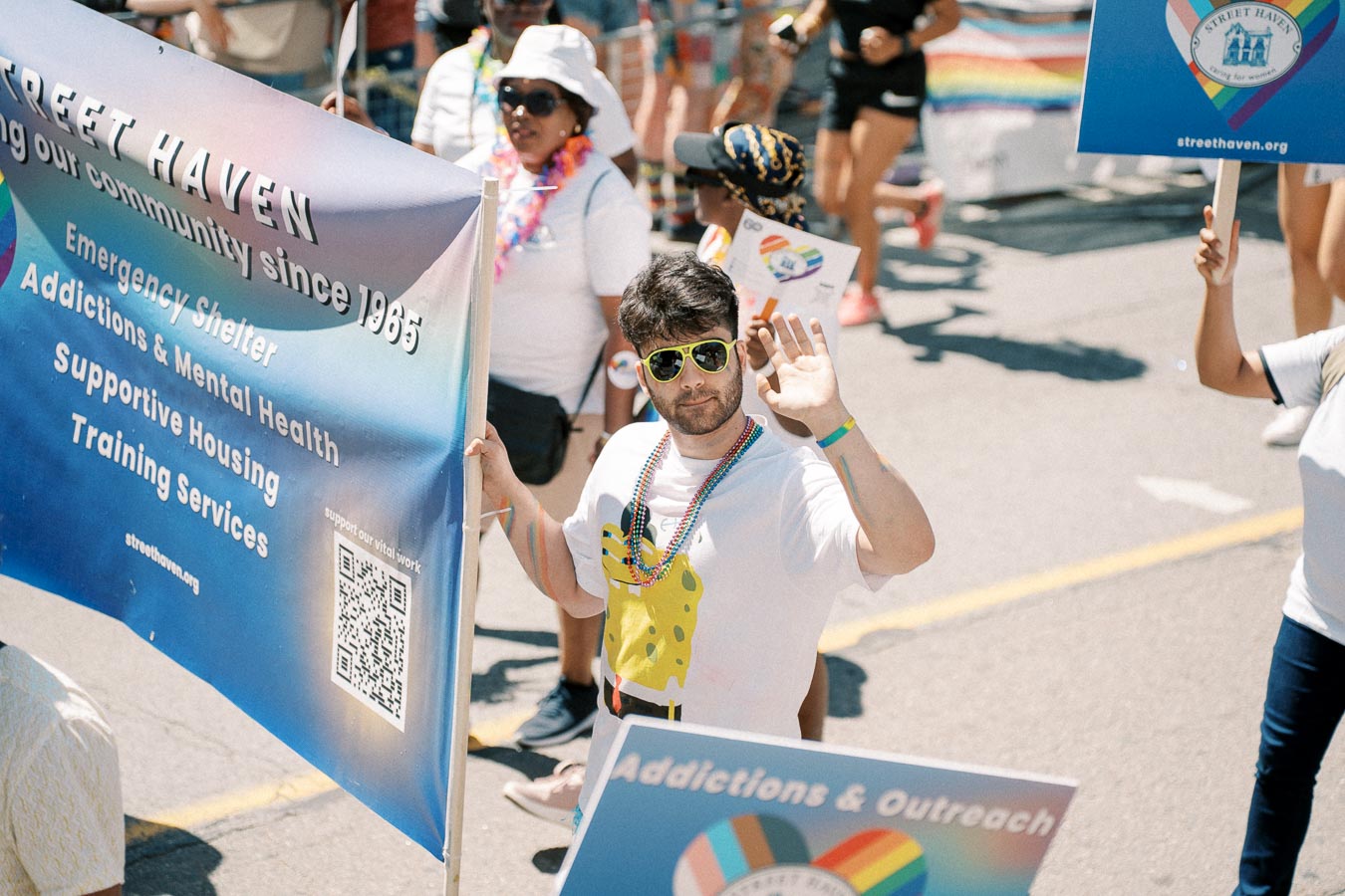 A participant waves while holding a 'Street Haven' banner at a parade, highlighting services like emergency shelter and mental health support.