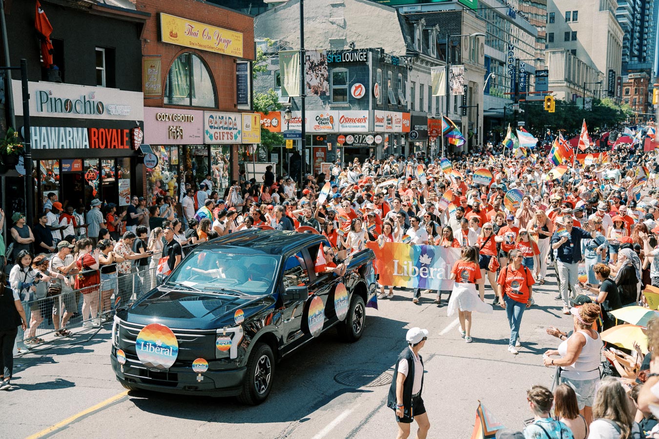 A lively parade celebrating diversity and inclusion, featuring a crowd with rainbow flags and banners, and a black truck adorned with rainbow stickers, moving through a bustling city street.