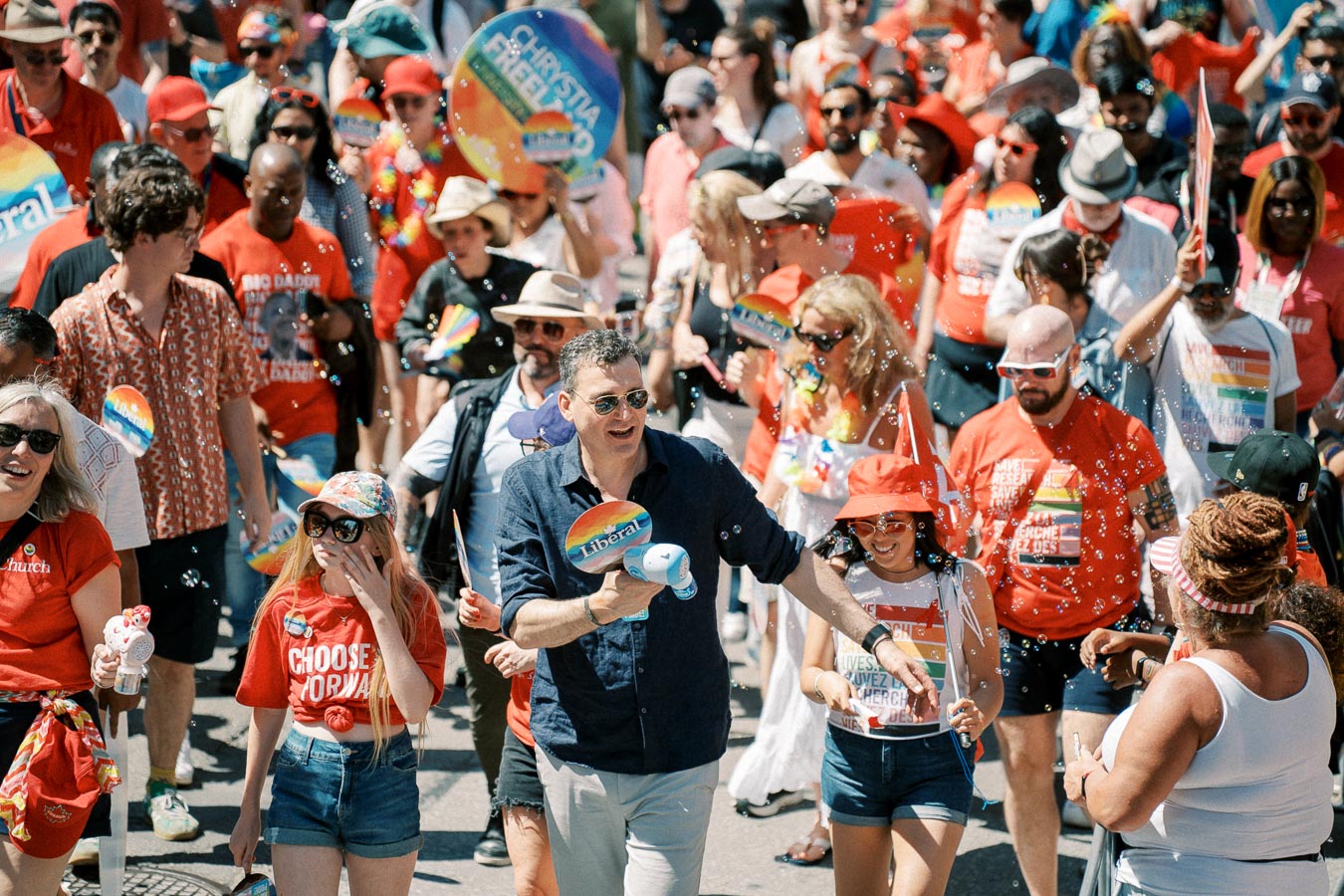 A lively parade scene with participants wearing red and rainbow-themed attire, celebrating with signs and banners.