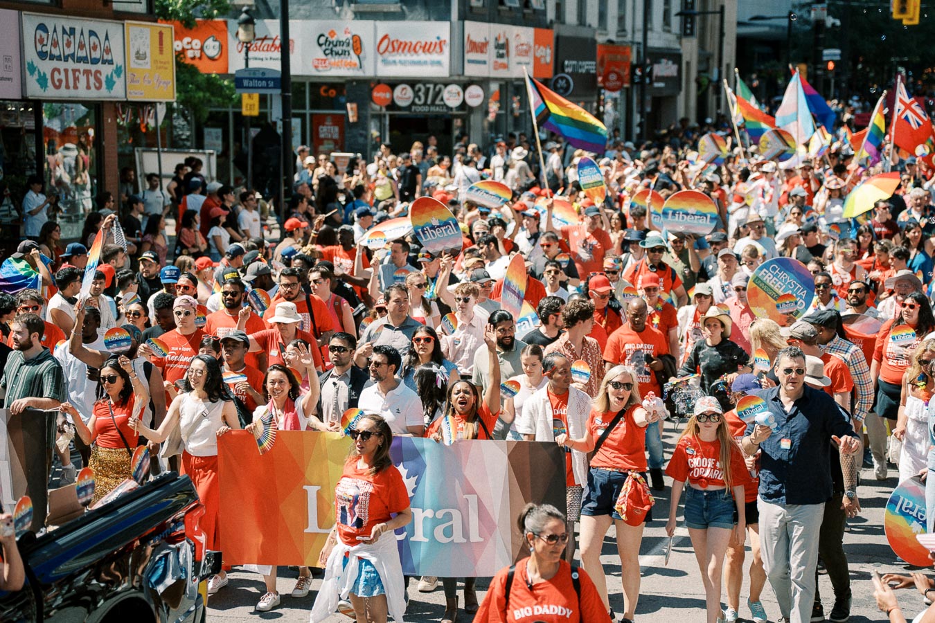 Participants in a parade hold rainbow flags and 'Liberal' signs, celebrating diversity and inclusion.