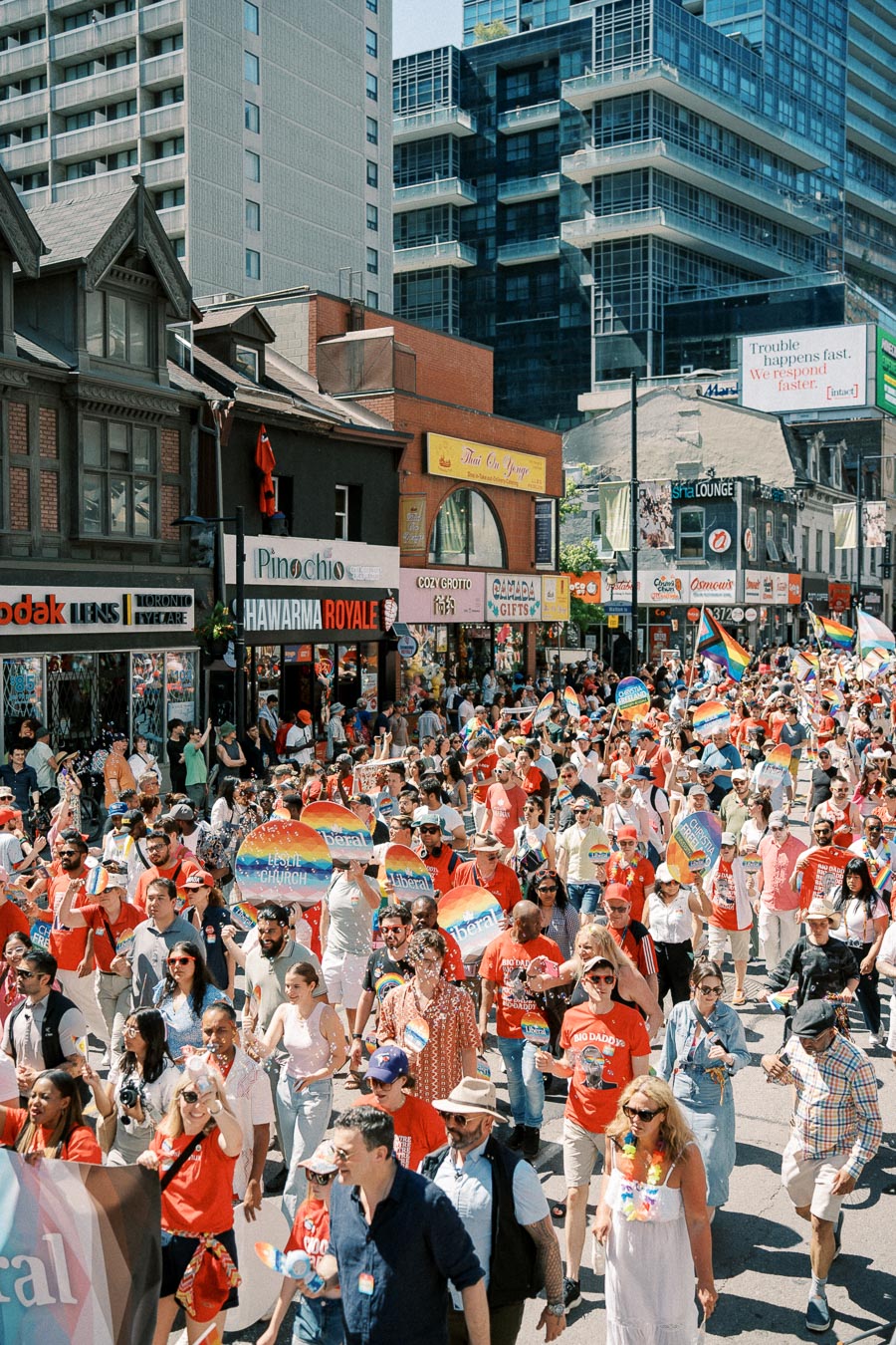A vibrant parade takes place on a city street, with participants celebrating and holding rainbow flags and signs. The street is bustling with people, and various shops and tall buildings are visible in the background.