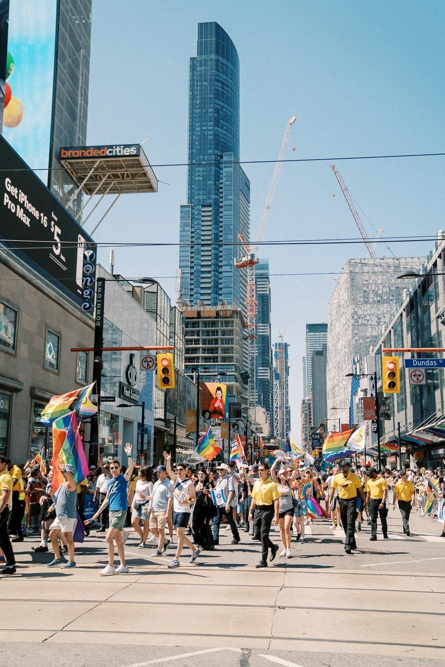 A lively parade featuring rainbow flags takes place on a city street, with skyscrapers and construction cranes visible in the background.
