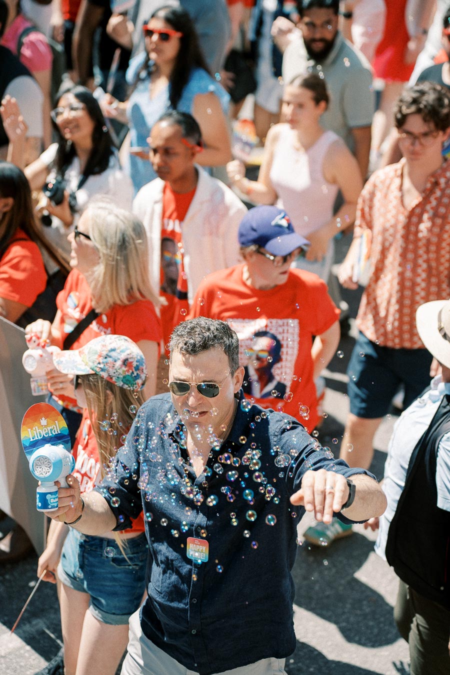 A lively scene at an outdoor event with a man blowing bubbles, surrounded by a crowd of people, many wearing red shirts.