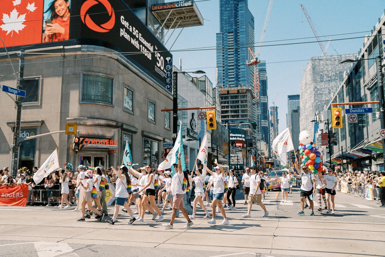 Participants march in a vibrant parade through a city intersection, holding flags and colorful balloons, with spectators lining the streets. The scene is set against a backdrop of skyscrapers and a large digital billboard.