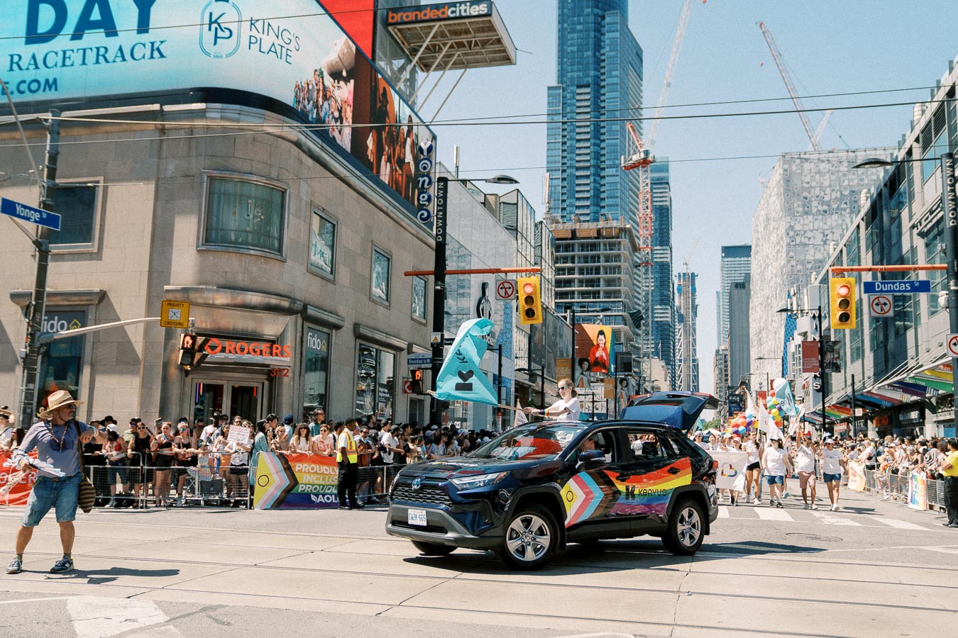 A vibrant parade takes place in a bustling city intersection, featuring a decorated SUV and participants carrying flags, with spectators lining the streets.