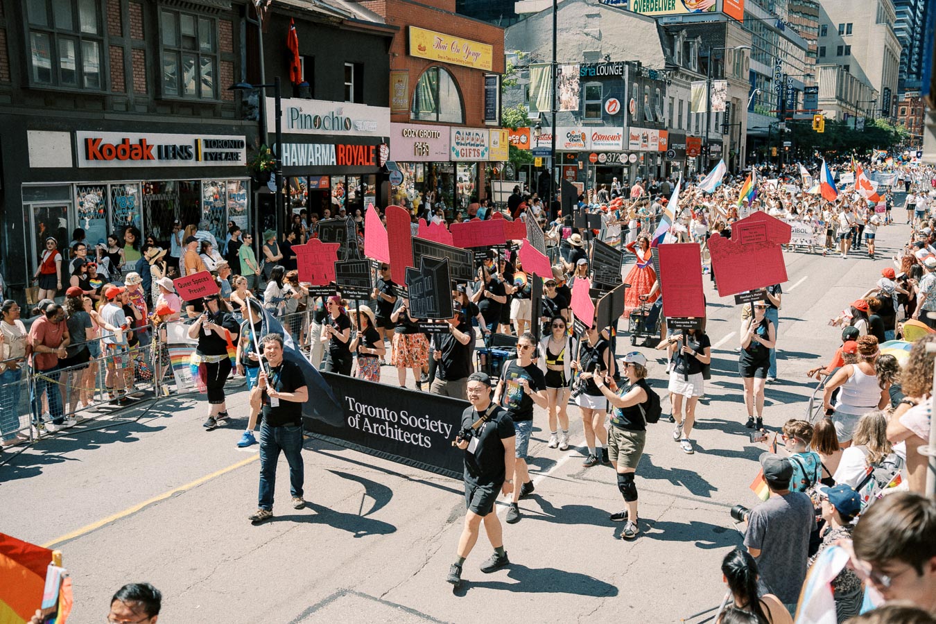 Participants from the Toronto Society of Architects march in a parade, holding signs and banners. The event takes place on a busy street in Toronto, with spectators lining the sidewalks and various storefronts in the background.