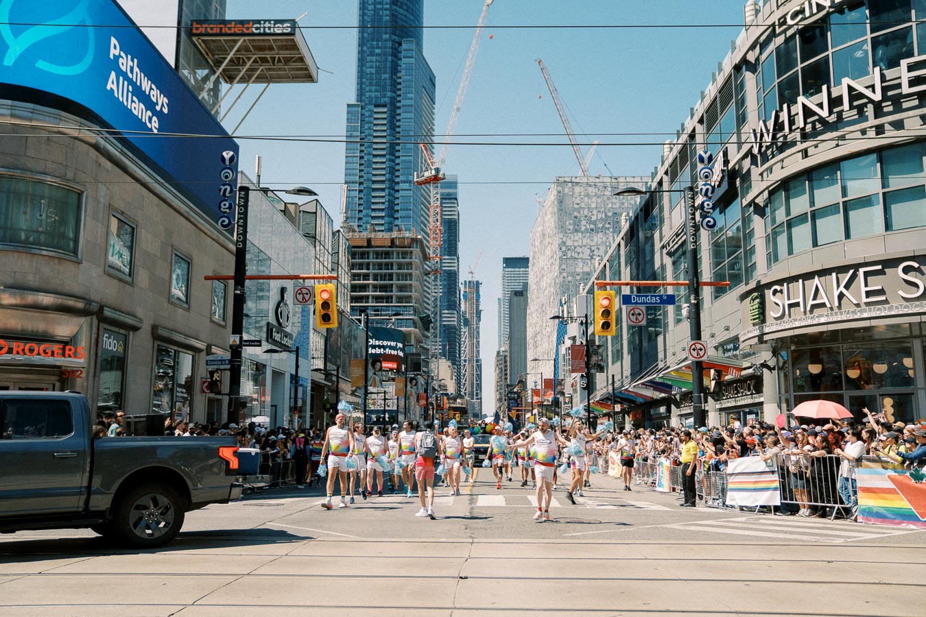 A lively parade takes place on Yonge Street in Toronto, with participants in colorful attire and a crowd of spectators. The scene is set against a backdrop of modern buildings and construction cranes.