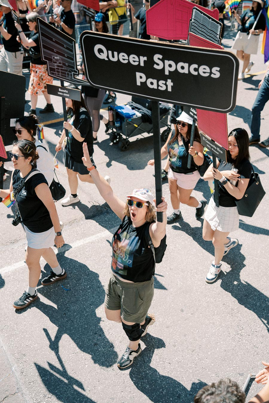 Participants in a parade celebrate with signs reading 'Queer Spaces Past', highlighting historical LGBTQ+ venues.