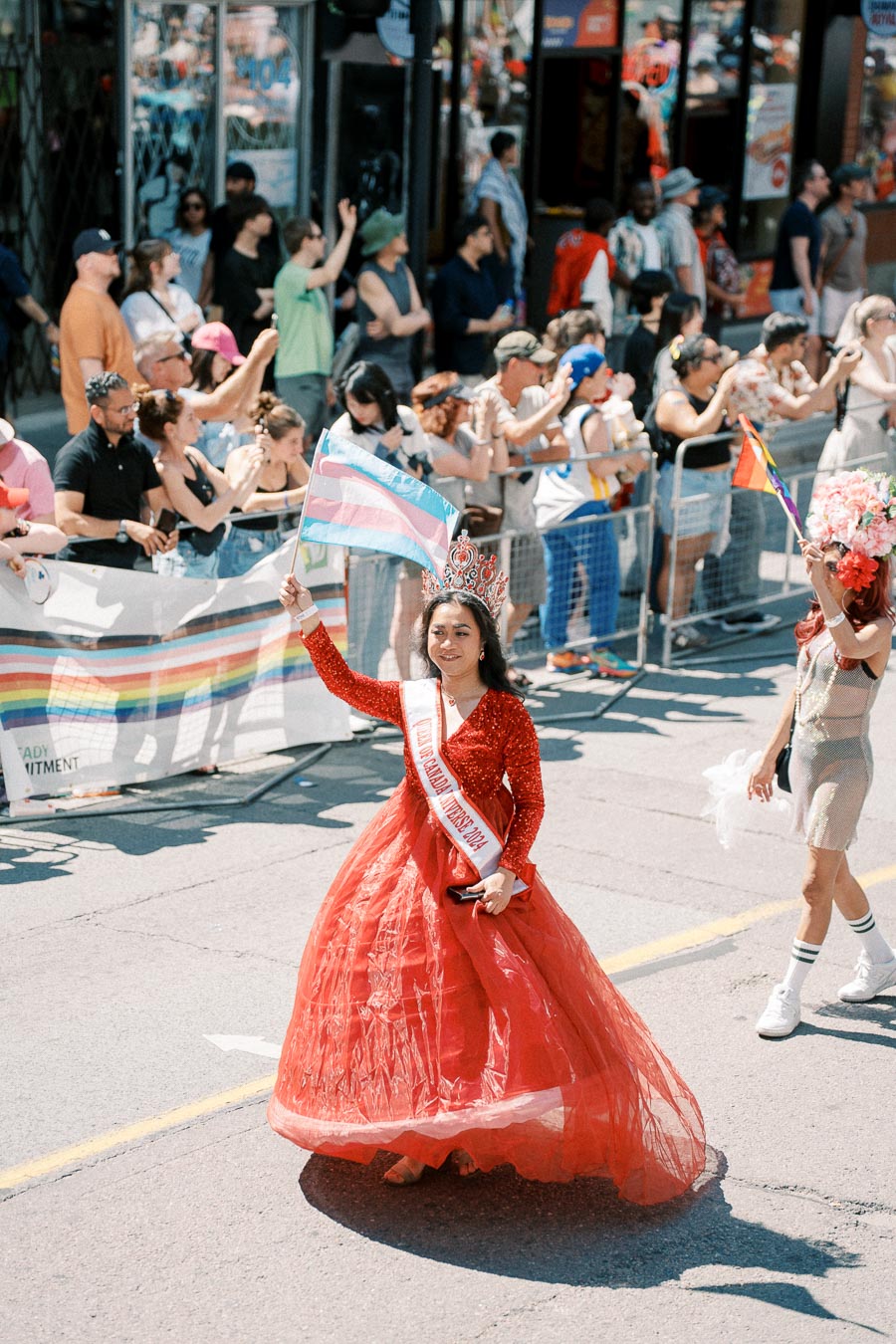 A parade participant in a red gown and crown waves a transgender pride flag, engaging with the crowd.