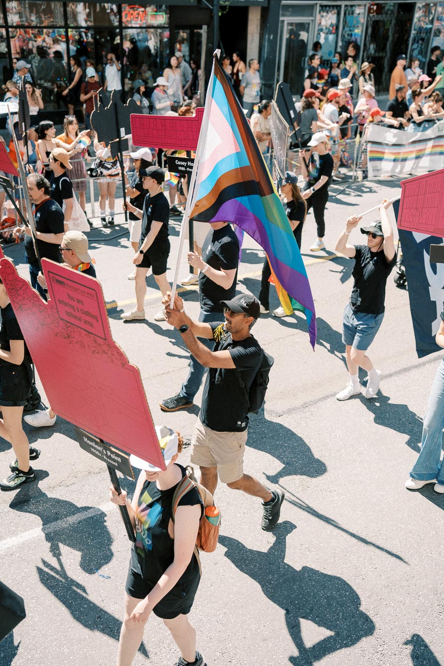 Participants in a pride parade march down the street, carrying flags and signs, while spectators watch from the sidelines.