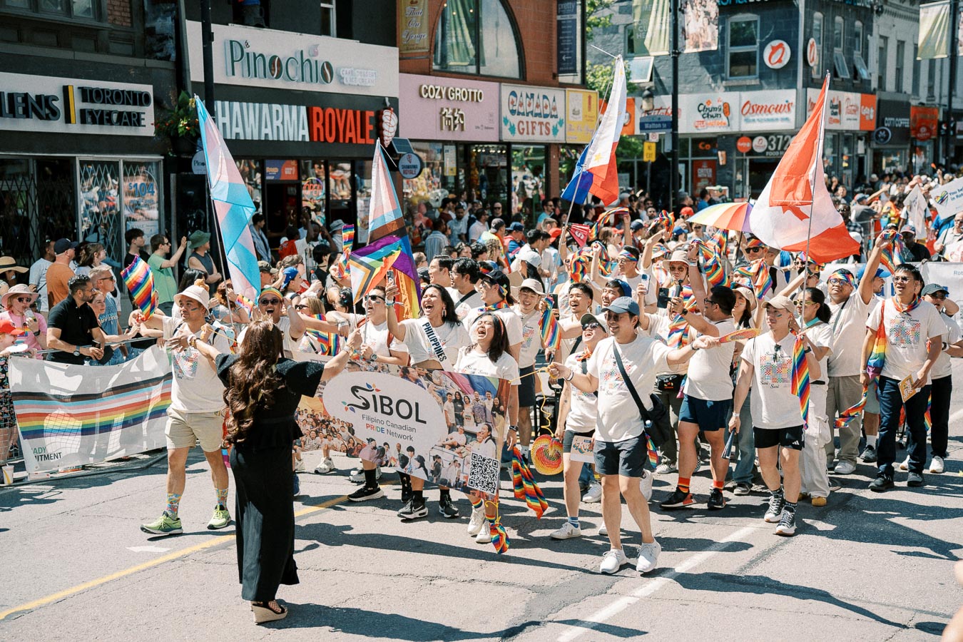Participants in a pride parade march joyfully, waving rainbow flags and banners, with a diverse crowd and storefronts in the background.