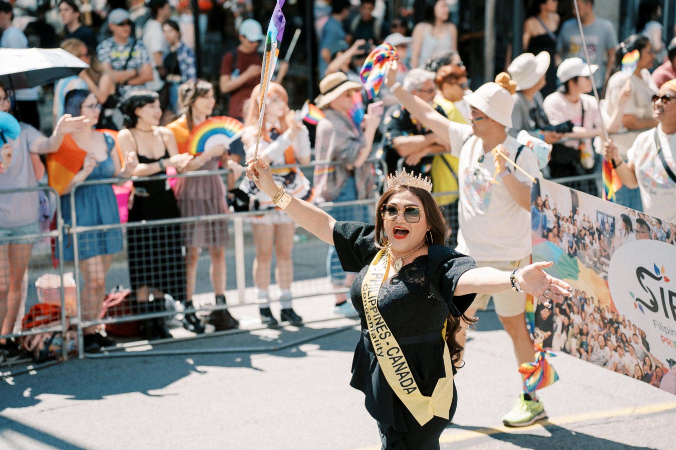 A participant in a pride parade joyfully waves a rainbow flag, surrounded by cheering spectators.