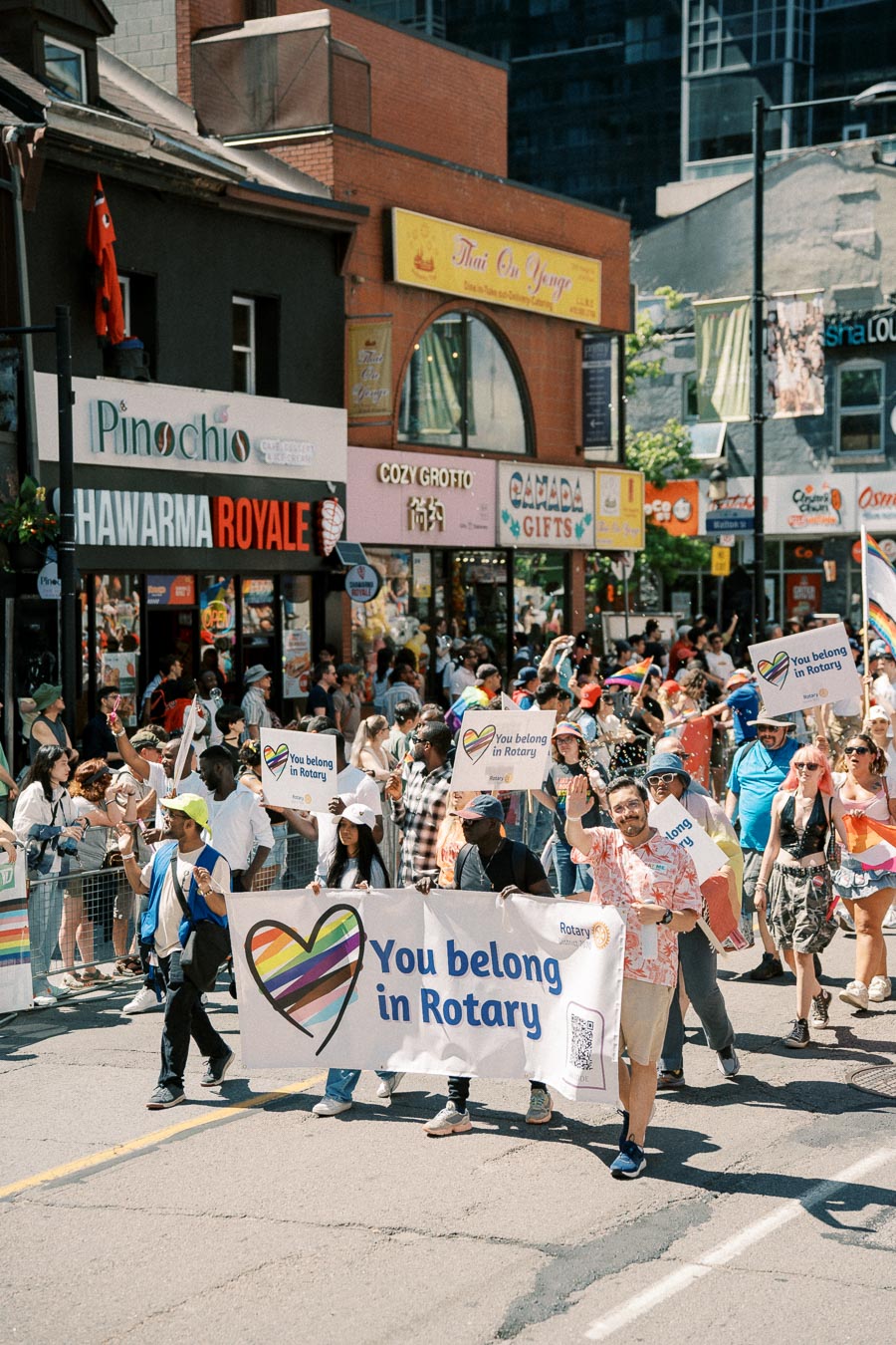 Participants in a parade hold a 'You belong in Rotary' banner, promoting inclusivity and diversity, as they march down a busy street lined with spectators and shops.