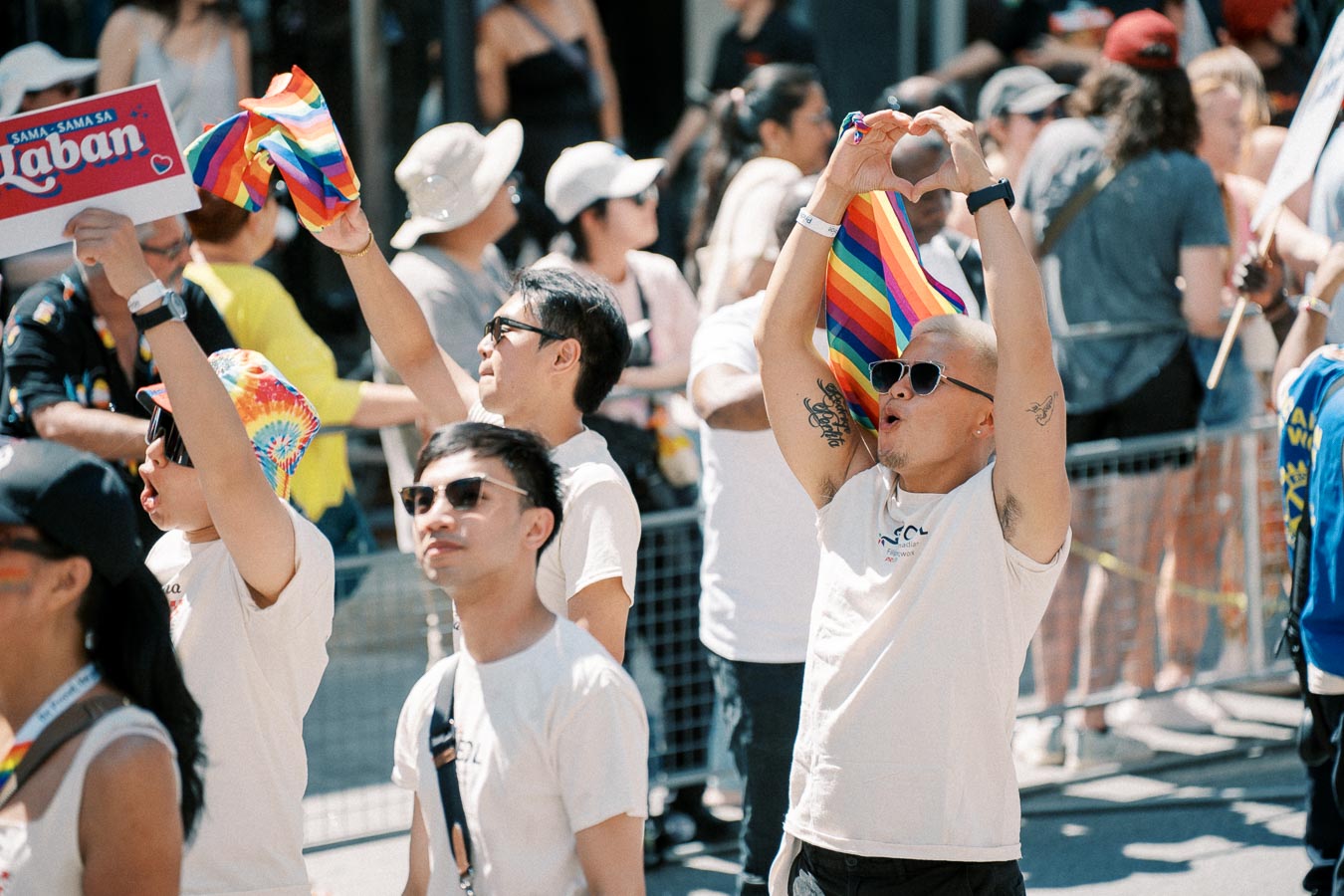 Participants celebrate at a pride parade, waving rainbow flags and signs, surrounded by a lively crowd.