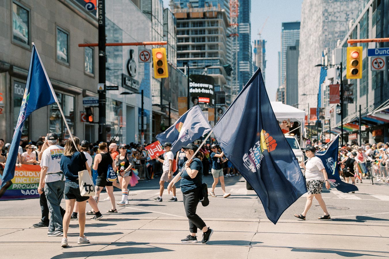 Participants in a pride parade march down a city street, carrying flags and banners, surrounded by a crowd of onlookers.