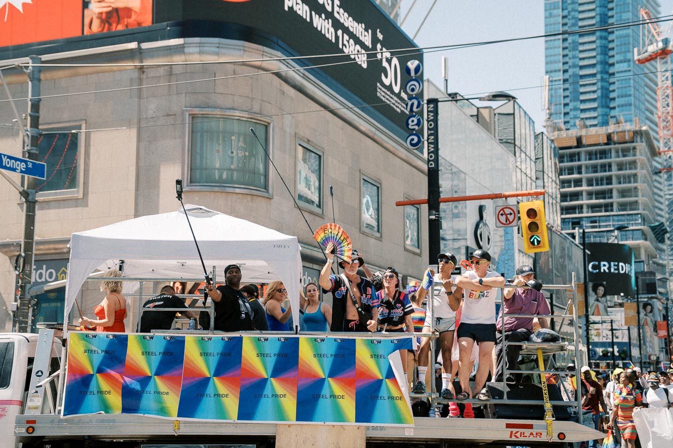 A vibrant parade float celebrating 'Steel Pride' with participants holding rainbow flags and banners, set against an urban backdrop.