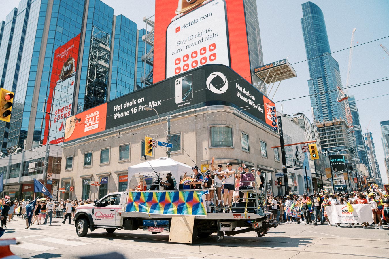 A vibrant parade float passes through a busy city intersection, surrounded by cheering crowds and towering skyscrapers. Large digital billboards display advertisements, adding to the lively urban atmosphere.