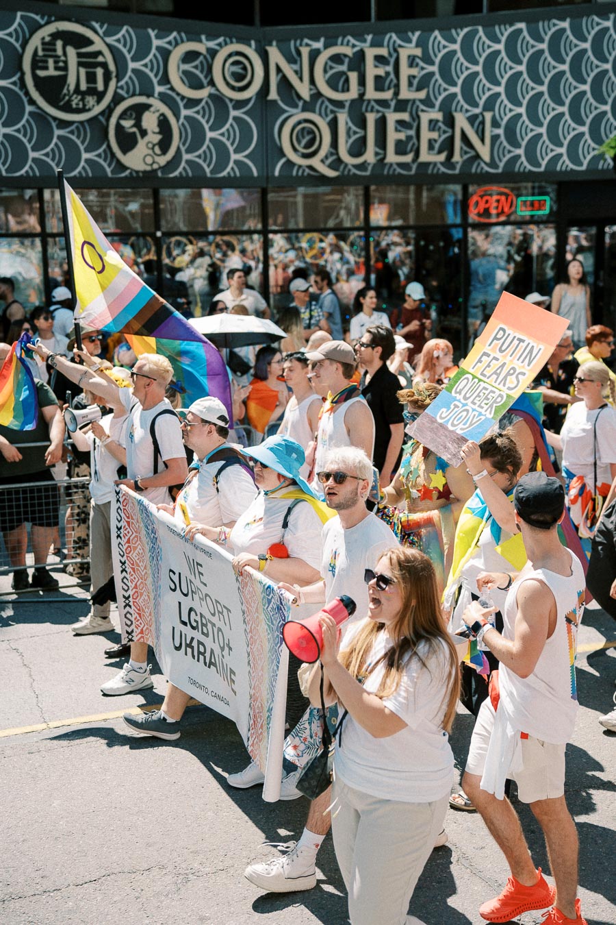 Participants in a pride parade march with signs supporting LGBTQ+ rights and Ukraine, in front of a restaurant named Congee Queen.