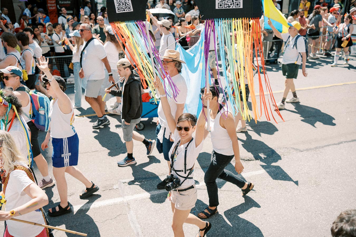 Participants in a vibrant parade hold rainbow streamers and signs with QR codes, celebrating diversity and inclusion.