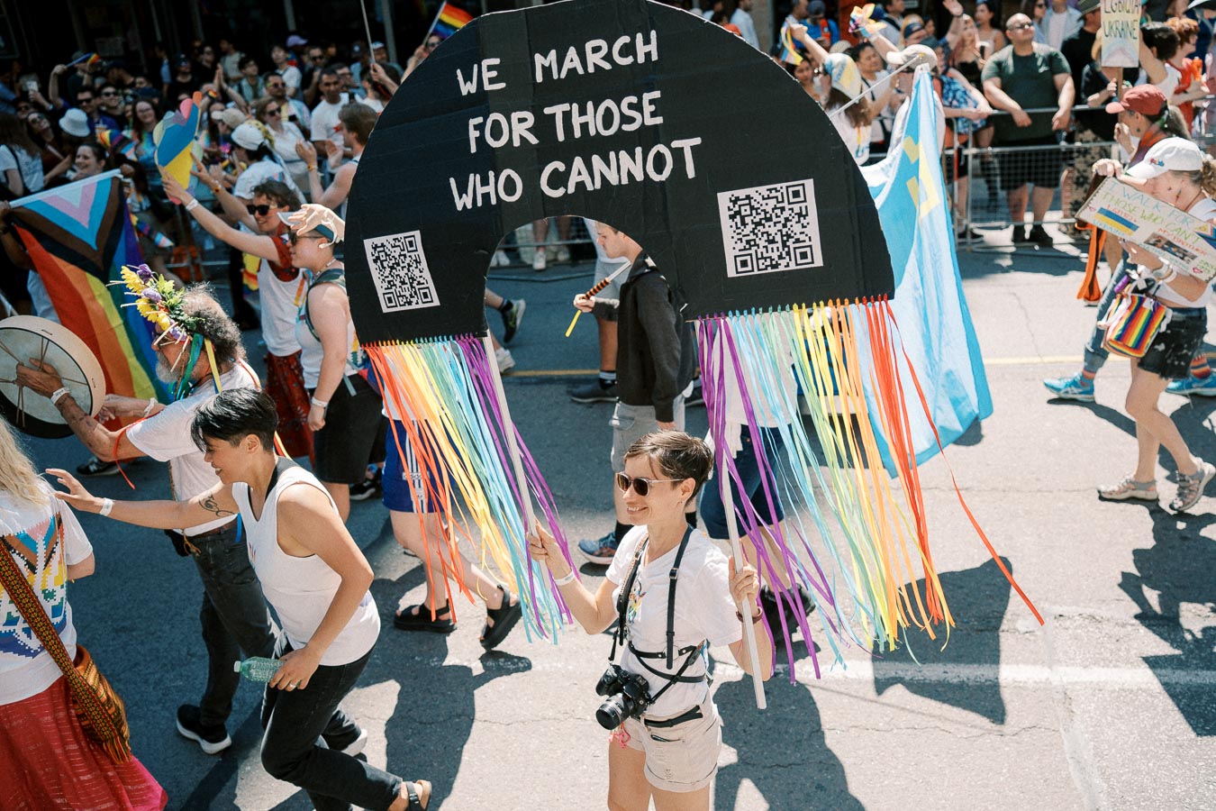 A participant at a pride parade holds a sign reading "We march for those who cannot," adorned with rainbow ribbons. The event is lively, with many people carrying flags and banners, celebrating diversity and inclusion.