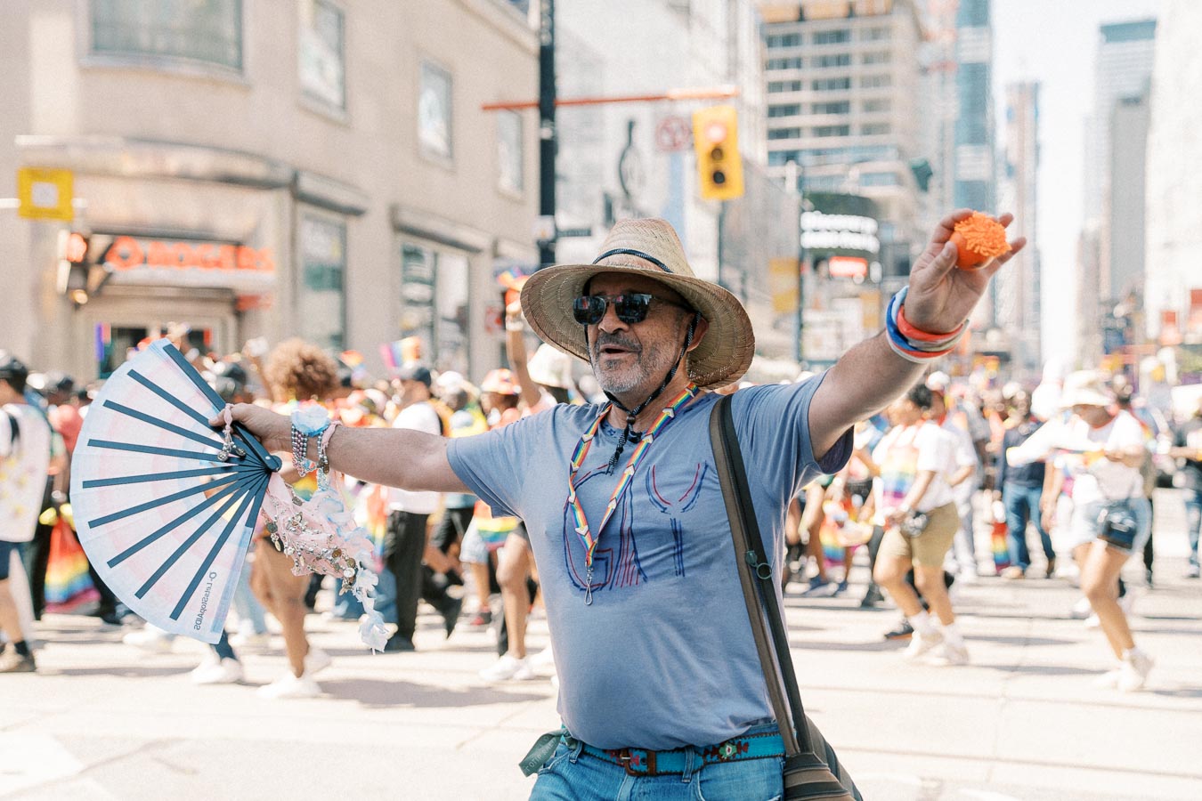 A lively scene from a pride parade featuring a participant in a straw hat and sunglasses, holding a colorful fan and wearing a rainbow necklace. The background is filled with a diverse crowd celebrating with rainbow flags and attire.