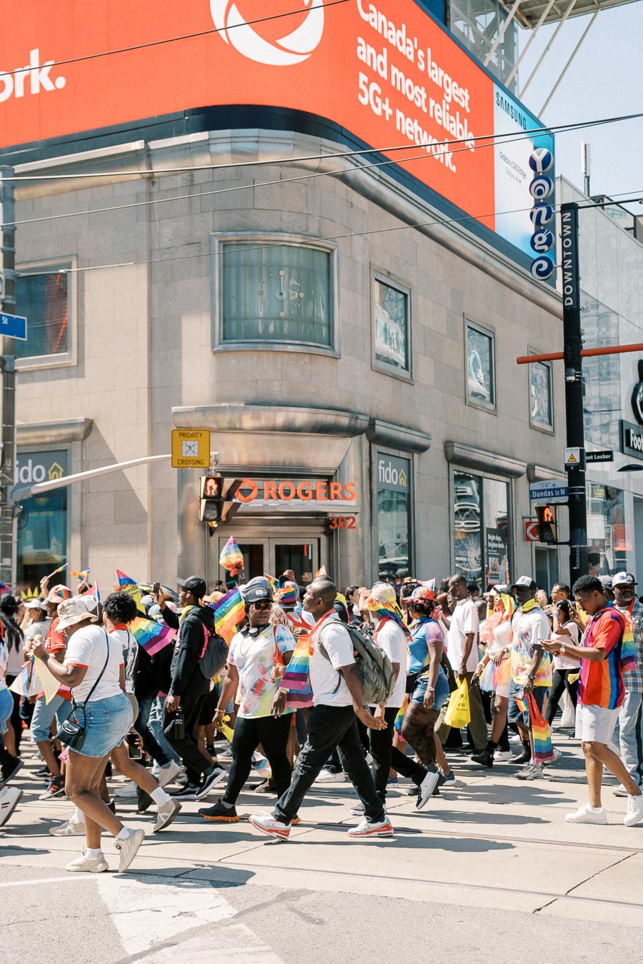 Participants in a pride parade walk past a Rogers store at a busy city intersection, holding rainbow flags and wearing colorful attire.