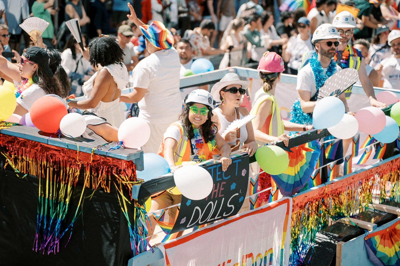 Participants on a colorful parade float celebrate with balloons and rainbow decorations, engaging with the crowd during a lively event.