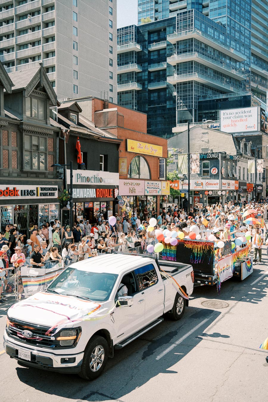 A vibrant parade featuring a white Ford truck and a colorful float passes through a bustling city street, lined with spectators and tall buildings.