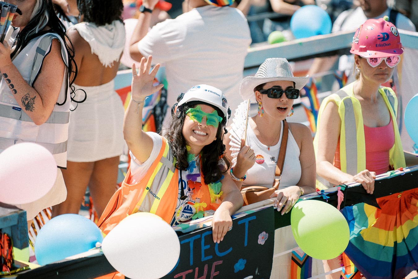 Participants at a pride parade wearing colorful outfits and hard hats, enjoying the festivities with balloons and rainbow decorations.