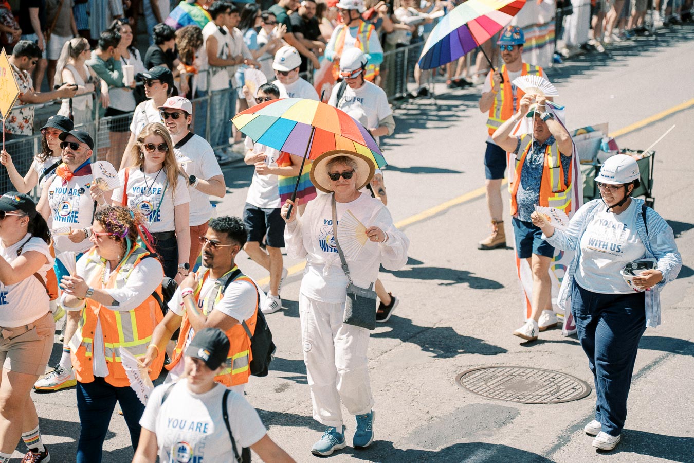 Participants in a parade walk down the street, some holding rainbow umbrellas and wearing colorful attire, while spectators watch from the sidelines.