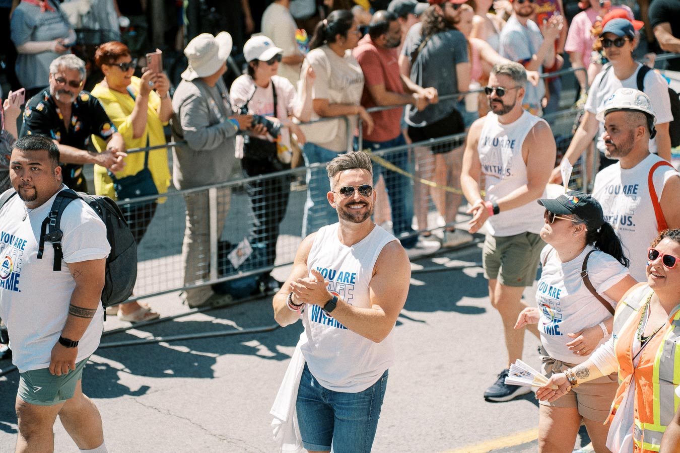Participants in a parade wearing matching shirts walk past spectators on a sunny day.