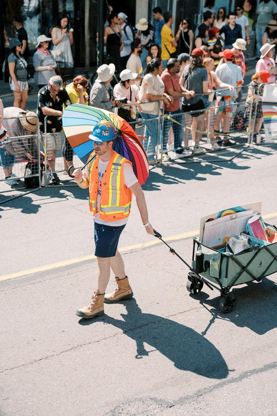 A parade participant wearing a construction vest and hard hat carries a rainbow umbrella and pulls a cart filled with signs, while a crowd watches from behind a barrier.