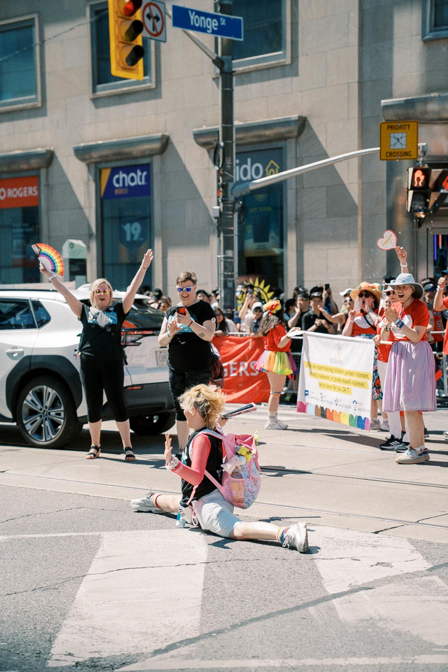 A lively parade scene on Yonge Street, featuring a person performing a split on the street while spectators cheer and hold a colorful banner.