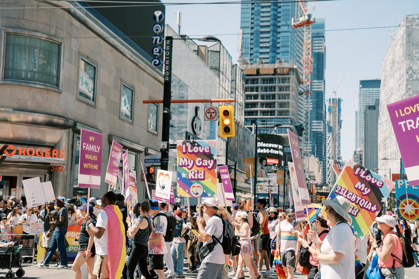 Participants in a pride parade march through a city street, holding signs supporting LGBTQ+ rights. The scene is lively with colorful banners and a diverse crowd.