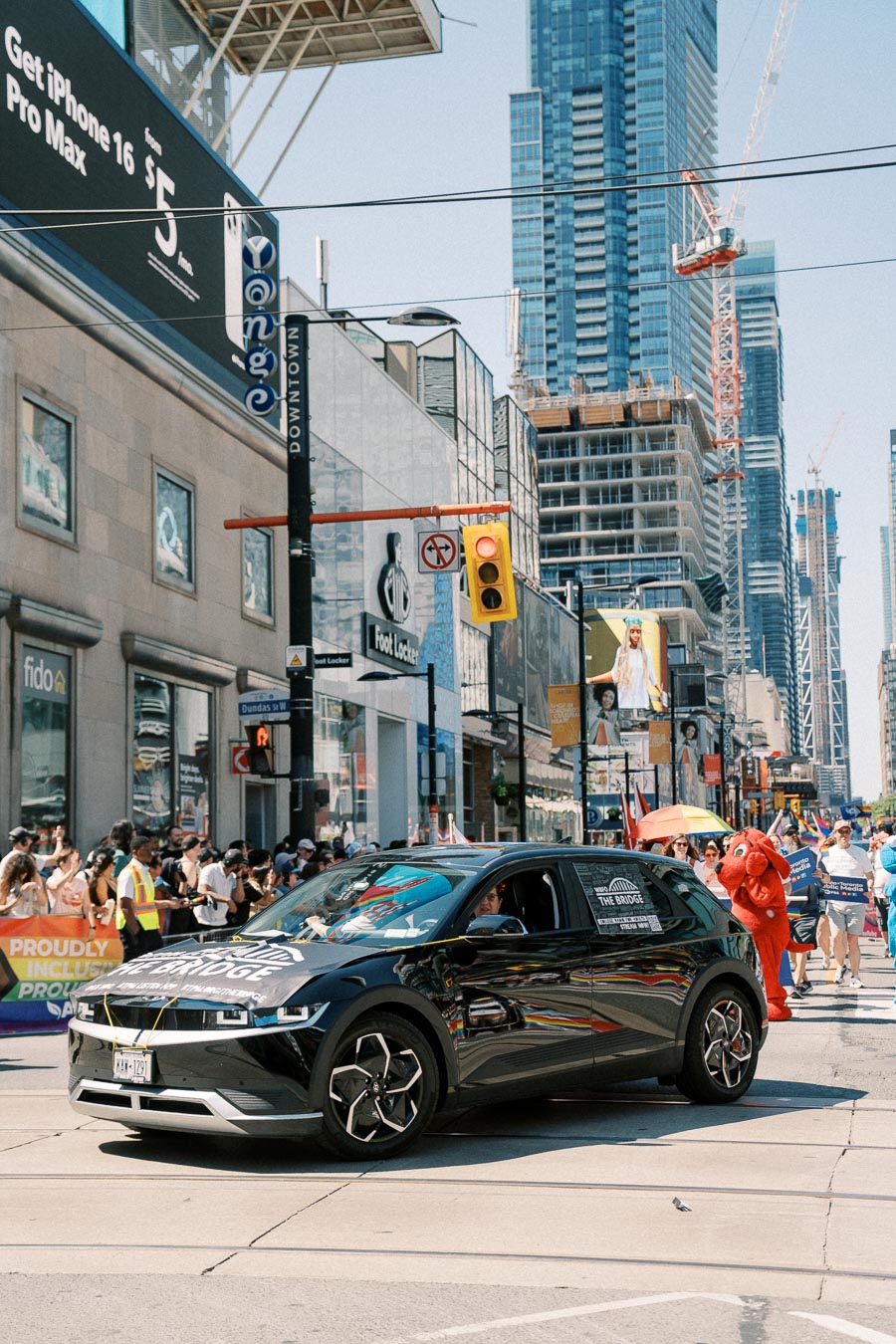 A black car participates in a city parade, surrounded by a crowd and skyscrapers in the background.