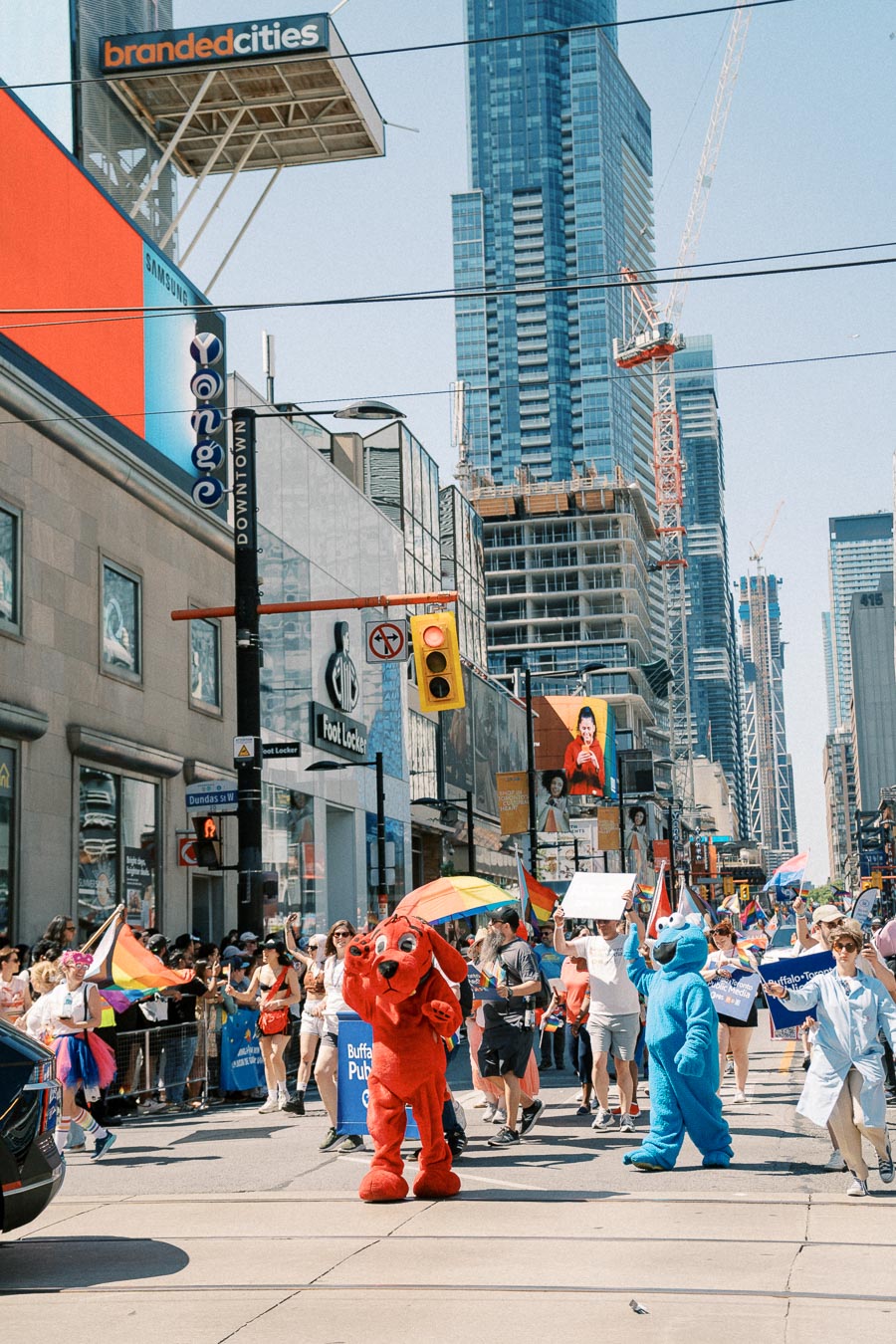 A lively parade in an urban setting with participants carrying rainbow flags and wearing colorful costumes, set against a backdrop of skyscrapers and construction cranes.