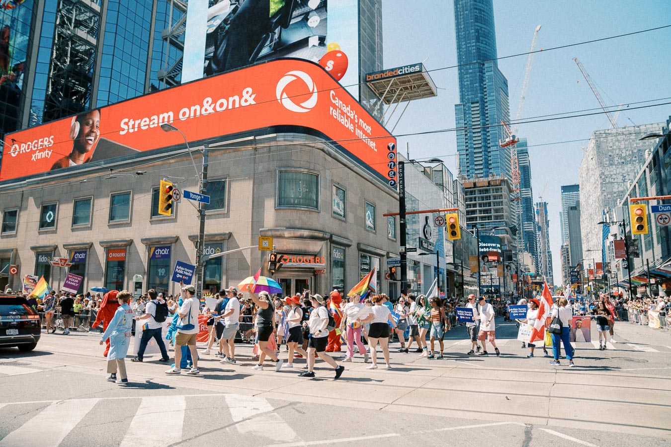 A vibrant parade takes place on a bustling city street, with participants carrying flags and banners. The scene is set against a backdrop of tall buildings and prominent advertisements, creating a lively urban atmosphere.