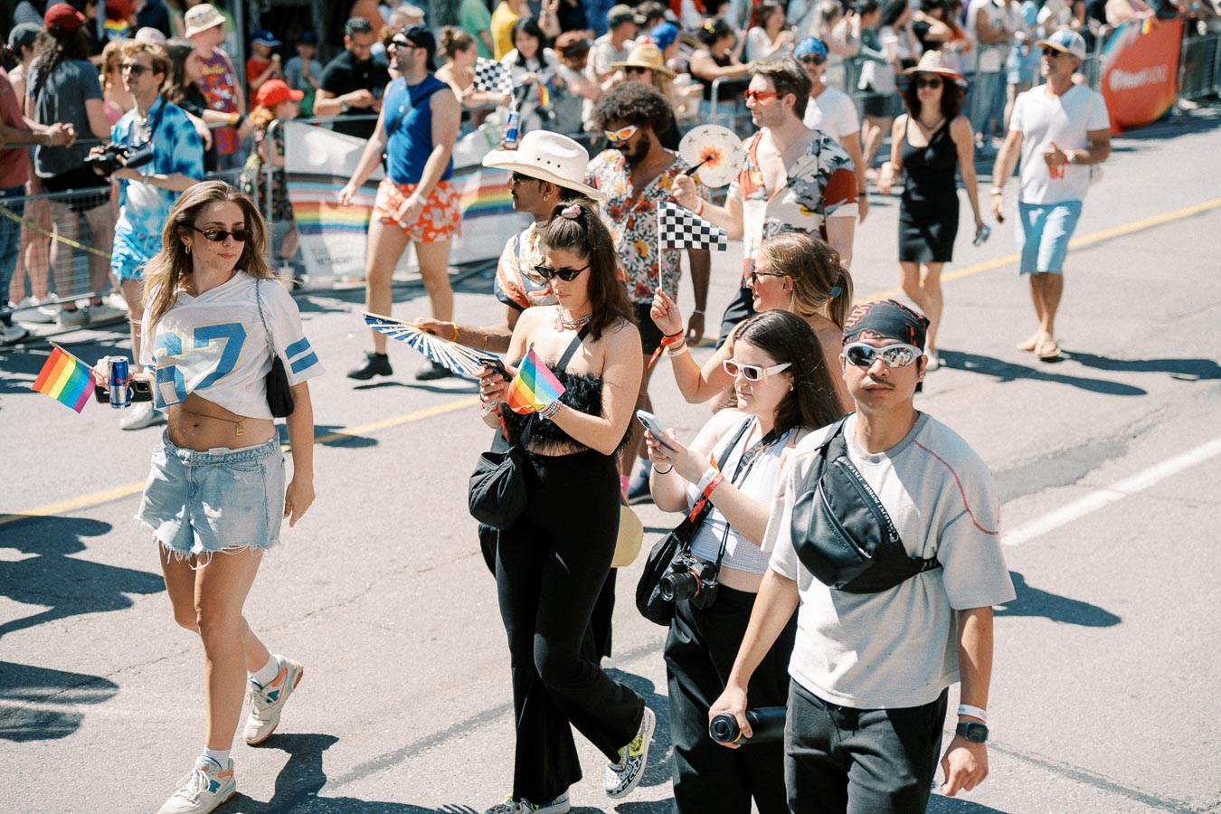 Participants in a pride parade walk down the street, holding rainbow flags and wearing casual summer attire. The crowd is lively, with people capturing moments on their phones and cameras.