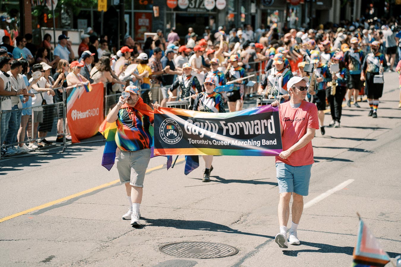 Participants in a parade hold a banner for 'Gayly Forward: Toronto's Queer Marching Band', celebrating with colorful attire and rainbow flags.
