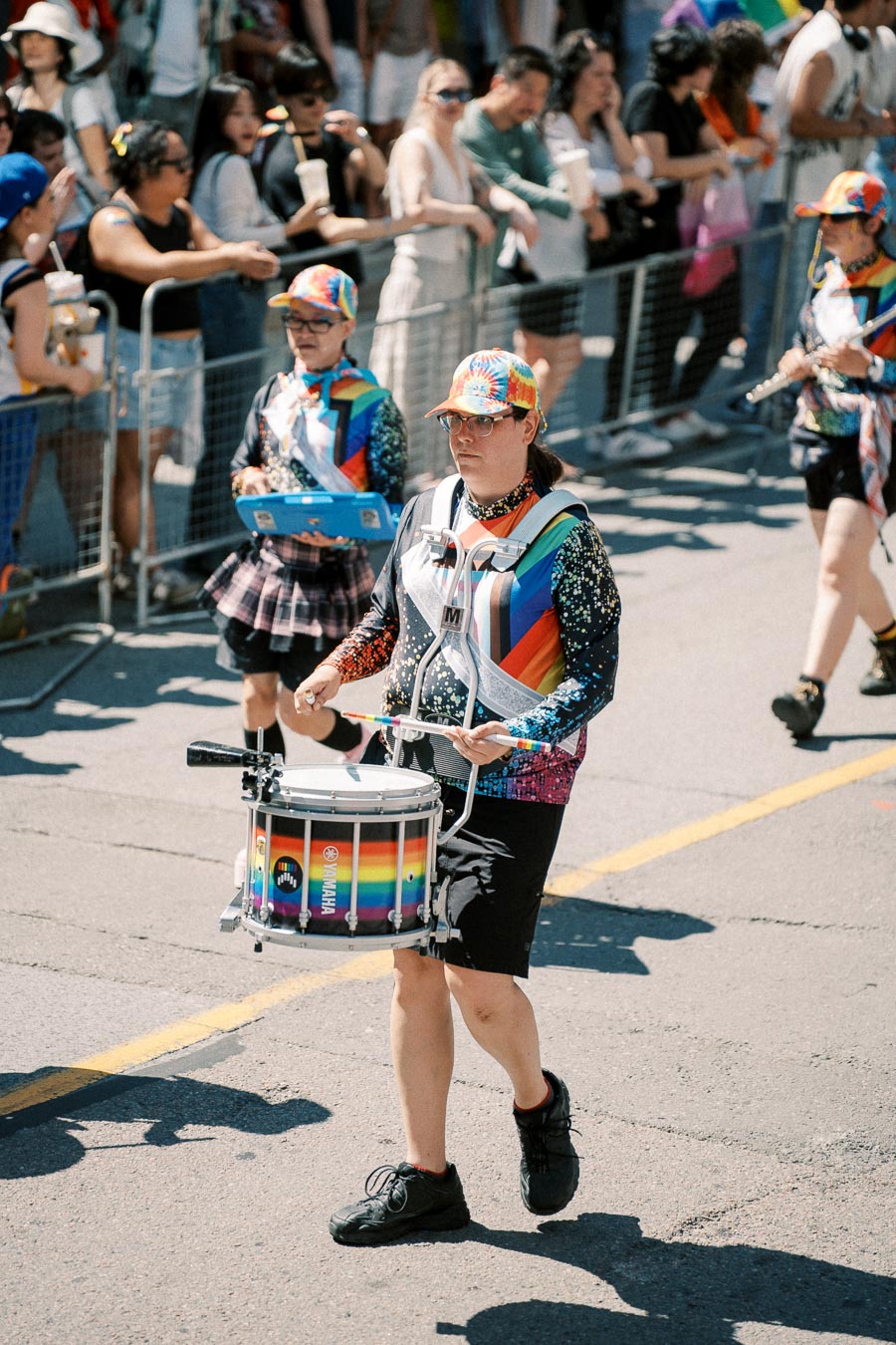 A parade participant in vibrant attire plays a drum adorned with rainbow colors, entertaining a crowd during a festive event.