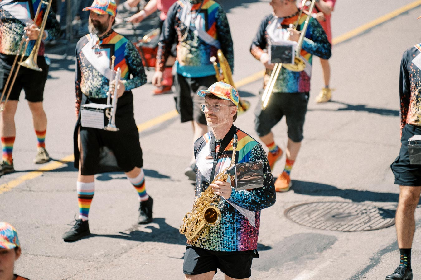 Musicians in a parade wearing vibrant, rainbow-themed outfits, playing instruments such as saxophones and trumpets.